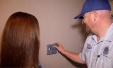 The image shows a man wearing a blue cap and a white shirt with a logo on it. He is standing in front of a beige wall and is...
