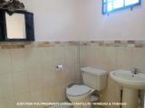 The image shows a small bathroom with a white toilet and a white sink. The walls are covered in beige tiles and there is a sm...