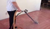 The image shows a person using a vacuum cleaner to clean a pink carpet. The person is wearing a white t-shirt and black pants...