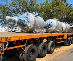 The image shows a large orange flatbed trailer with four large cylindrical tanks attached to it. The tanks are wrapped in pla...