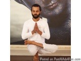 The image shows a man sitting in a yoga pose with his hands clasped together in a meditative manner. He is wearing a white tr...
