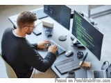 The image shows a man sitting at a desk in front of two computer monitors. He is wearing a black sweater and glasses and appe...