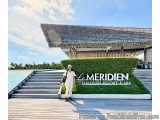 The image shows a woman standing in front of a large sign that reads "Le Meridien Maldives Resort & Spa". The sign is located...