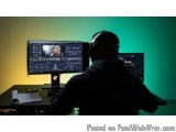 The image shows a person sitting at a desk in front of two computer monitors. The person is wearing a black jacket and has a...