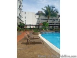 The image shows a large swimming pool with crystal clear blue water. There are two lounge chairs on the left side of the pool...