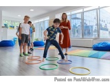 The image shows a group of children and a woman in a room with large windows. The children are playing with colorful hula hoo...