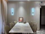 The image shows a dining room with a long white table in the center. The table is surrounded by six beige chairs. On the tabl...