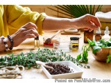 The image shows a person's hands holding a small bottle of essential oil and applying it to a small bowl of herbs. The person...