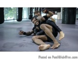 The image shows two men engaged in a mixed martial arts (MMA) fight in a gym. The man on the left is lying on the ground with...