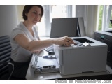 The image shows a woman sitting at a desk in an office. She is wearing a white t-shirt and is focused on the printer in front...