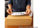 The image shows a person's hands holding a piece of paper with a barcode on it. The person is standing in front of a cardboar...
