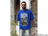 The image shows a man standing in front of a stone wall with a wooden door. He is wearing a blue t-shirt with an illustration...