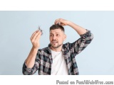 The image shows a young man with a beard and mustache, wearing a plaid shirt. He is standing against a light blue background...
