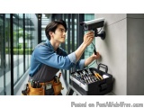 The image shows a young man wearing a blue shirt and a tool belt with various tools in it. He is standing in front of a gray...