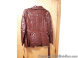 The image shows a maroon leather jacket hanging on a wooden door. The jacket has a high collar and long sleeves. It has a bel...
