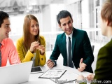 The image shows a group of four people, two men and two women, sitting around a table in a meeting room. They are engaged in...