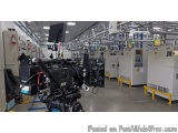 The image shows a large industrial space with a large camera mounted on a tripod in the center. The camera is black and appea...
