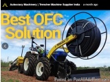 The image shows a blue and yellow tractor with a large spool of black hose attached to it. The tractor is parked on a dirt fi...