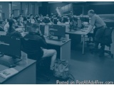 The image is a black and white photograph of a large room with multiple computer desks and chairs. The desks are arranged in...