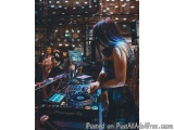 The image shows a young woman in a black dress playing a DJ set at a nightclub. She is standing in front of a Pioneer DJ mixe...
