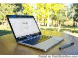The image shows a laptop computer sitting on a wooden table in a park. The laptop is open and the screen is turned on, displa...