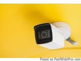 The image shows a white security camera mounted on a yellow wall. The camera is round and has a black lens in the center. It...