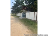 The image shows a dirt road leading to a gate with a wooden roof. The gate is made of metal and has a blue gate in the center...