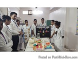 The image shows a group of young people in a classroom setting. They are all wearing white lab coats and are gathered around...