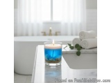 The image shows a glass candle on a white marble countertop in a bathroom. The candle is lit and has a blue liquid inside, wh...
