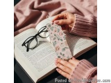 The image shows a pair of hands holding a bookmark with a floral design on it. The bookmark is resting on top of an open book...
