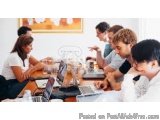 The image shows a group of six people sitting around a wooden table with laptops and papers scattered across it. They appear...