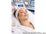The image shows a young woman lying on a white towel on her head with her eyes closed, receiving a facial treatment. She is w...