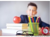 The image shows a young boy sitting at a desk with a stack of books, an apple, and a red alarm clock. He is wearing a blue sh...