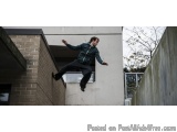 The image shows a young man in mid-air, performing a skateboard trick. He is wearing a green jacket, black pants, and black s...