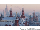 The image is a panoramic view of the Moscow Kremlin in Russia. The sky is a hazy pink color, indicating that it is either sun...