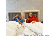 The image shows two elderly men lying on a bed with white sheets and pillows. They are both smiling and making a peace sign w...