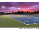 The image shows a tennis court at sunset. The sky is a beautiful mix of pink, purple, and orange hues, with the sun setting i...