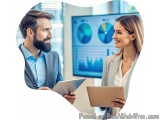 The image shows a man and a woman in an office setting, engaged in a conversation. They are both holding a tablet and appear...