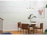 The image shows a modern dining room with a white brick wall and wooden flooring. There is a round wooden table with a vase o...