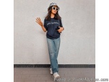 The image shows a young woman standing in front of a white wall. She is wearing a black t-shirt with the words "Adidas, bitch...