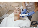 The image shows a young woman lying on a massage table with her eyes closed, receiving a facial treatment. She is wearing a w...