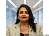 The image is a portrait of a middle-aged woman with shoulder-length dark hair. She is wearing a light beige blazer and a blac...