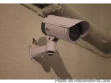 The image shows a security camera mounted on a white wall. The camera is round and has a white body with a green lens. It is...