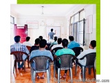 The image shows a classroom with a group of students sitting on chairs facing a whiteboard. The students are of different age...