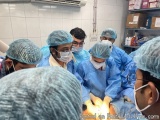 The image shows a group of surgeons performing an operation in an operating room. There are six surgeons in the image, all we...