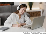 The image shows a young woman sitting at a desk with a laptop and a notebook in front of her. She is wearing a white blouse a...