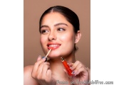The image is a close-up portrait of a young woman applying lipstick. She is holding a tube of lipstick in her right hand and...