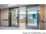 The image shows a modern entrance door in a hospital corridor. The door is made of stainless steel and has a glass panel on t...