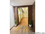 The image shows the entrance of a modern building with a wooden door. The door is framed by a dark brown wall and has a glass...