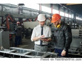 The image shows two men in a factory setting. They are both wearing hard hats and are standing in front of a large machine. T...